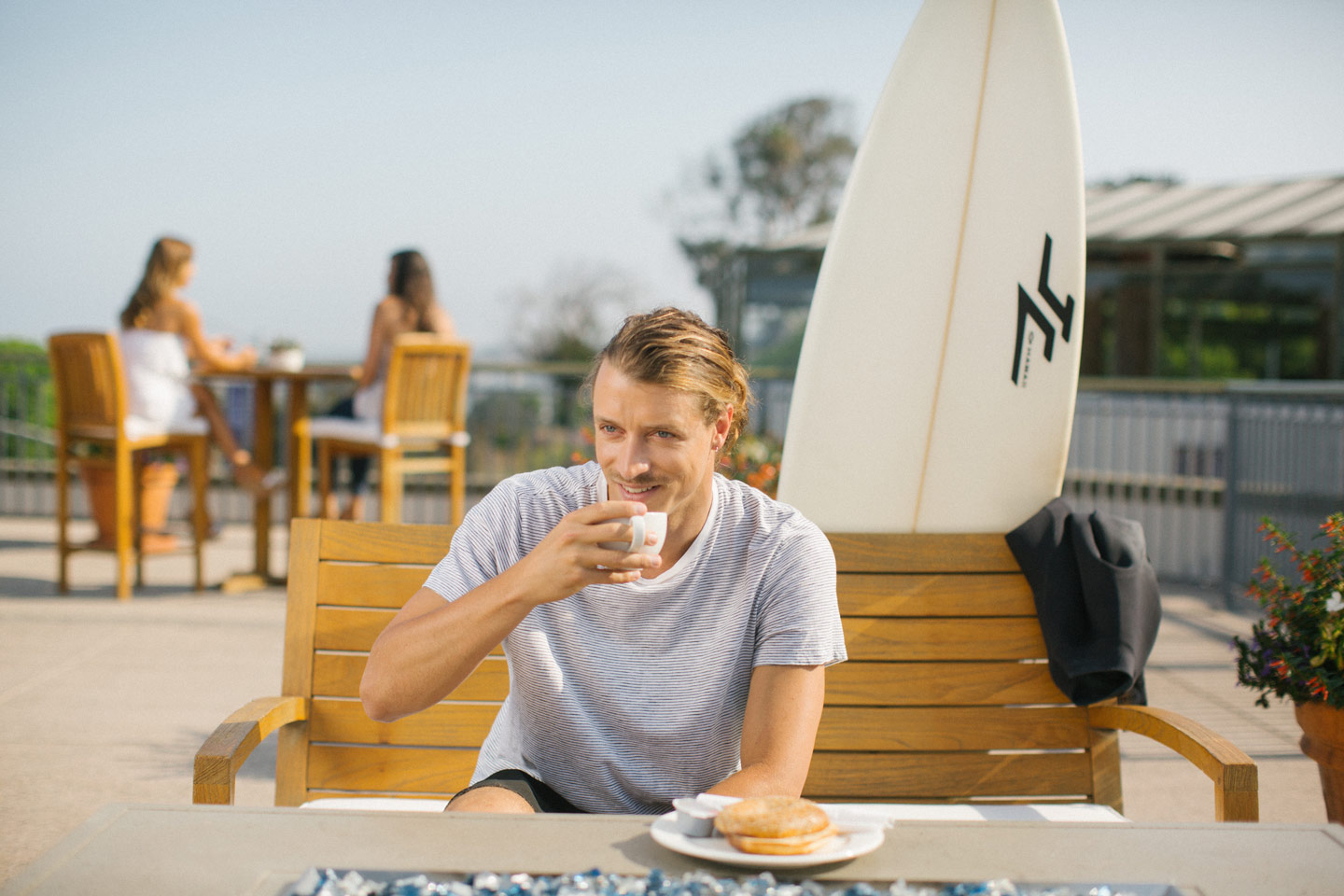 man sitting on bench in Del Mar Plaza, enjoying a drink and food. in the background is a blue sky, his surfboard, and some friends at another table
